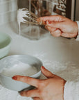A woman holding a gold whisk while it drips white vanilla frosting into a blue bowl.