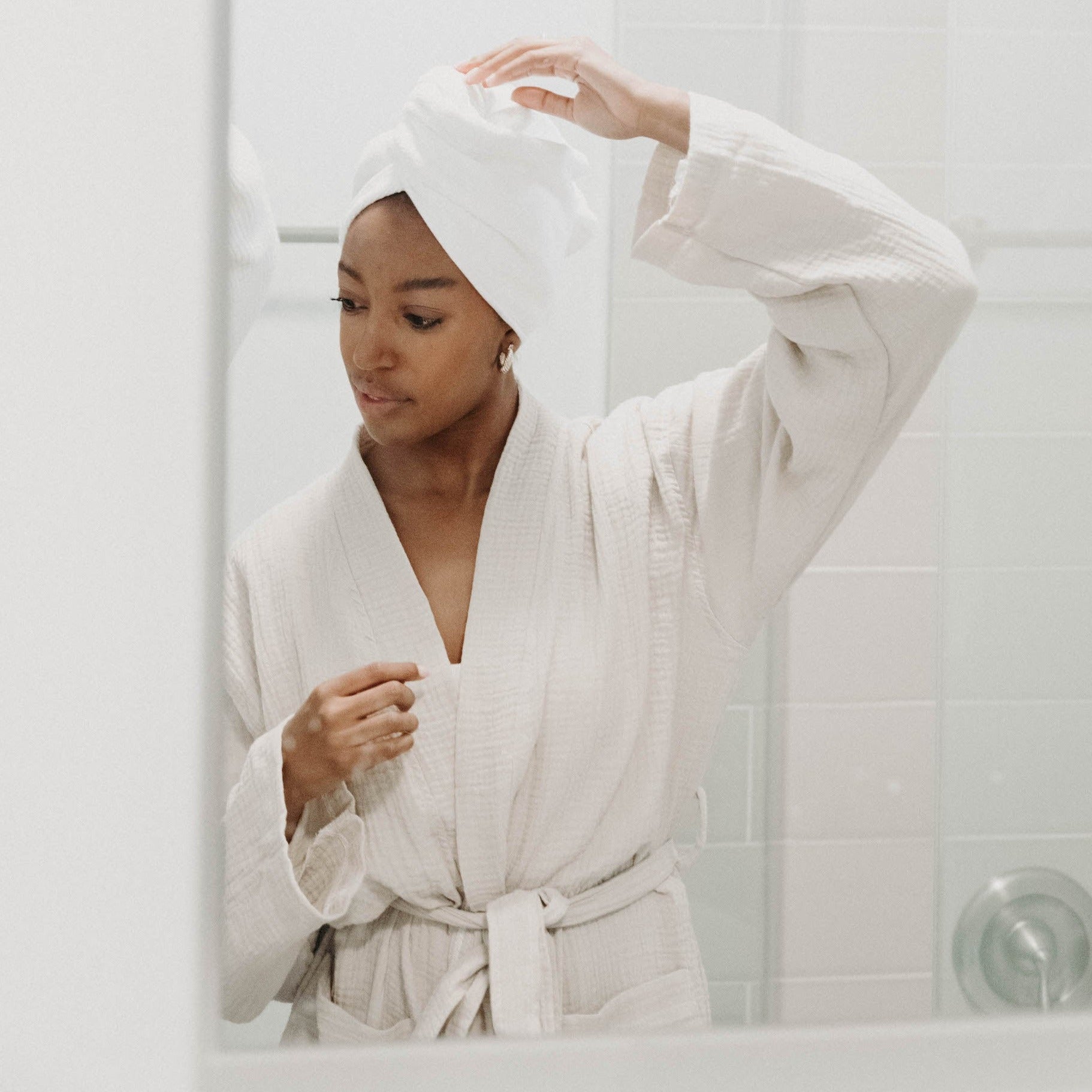 A girl standing in a bathroom mirror wearing a muslin cloud oatmeal robe with a better hair days ahead hair towel in her hair.