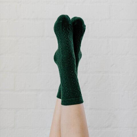 A pair of legs raised against a light beige wall, wearing dark green mid-calf socks with a textured knit pattern featuring diagonal lines and diamond shapes.