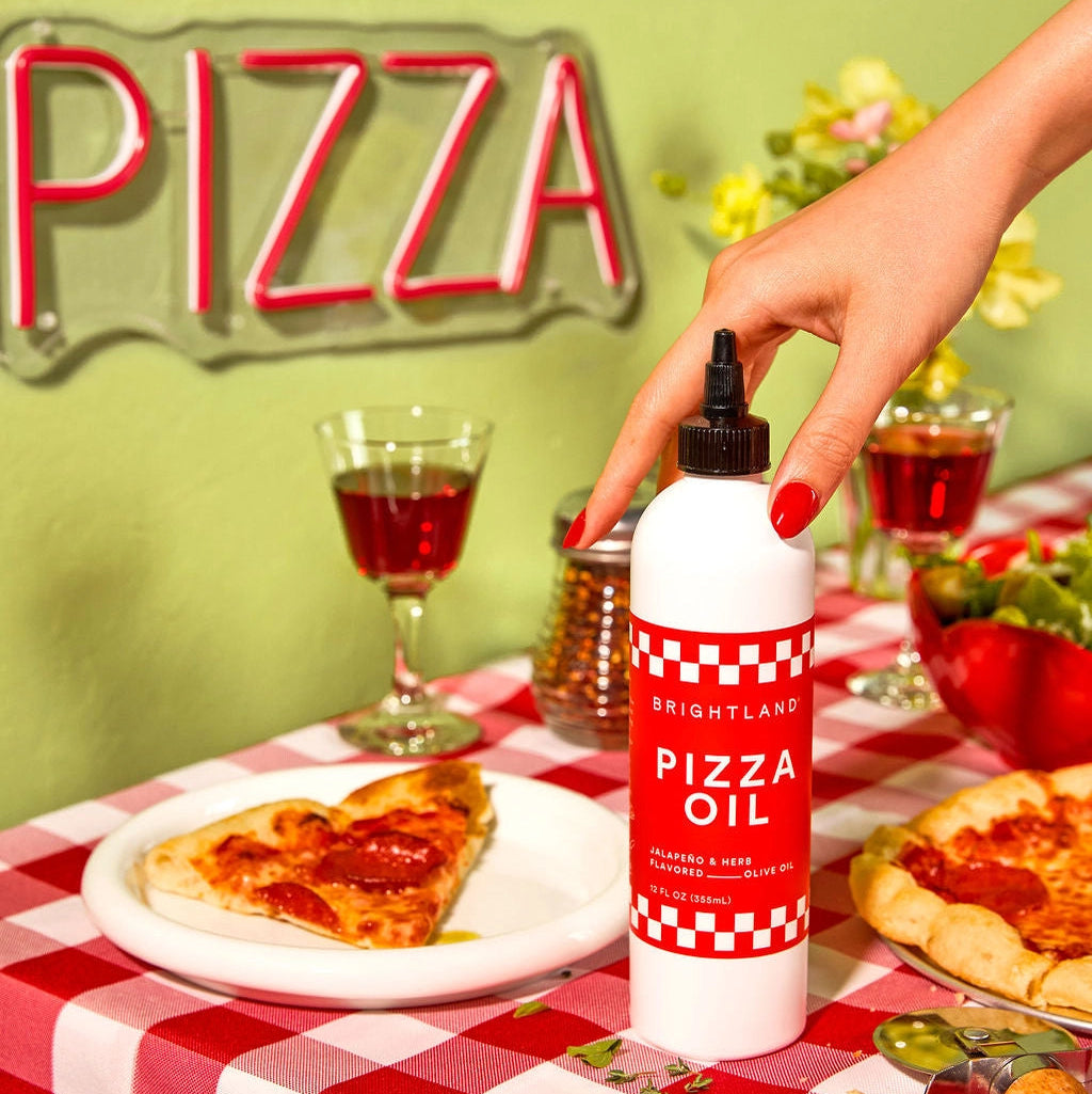 Person holding a bottle of pizza oil over a pizza on a checkered tablecloth with a 'PIZZA' sign in the background.