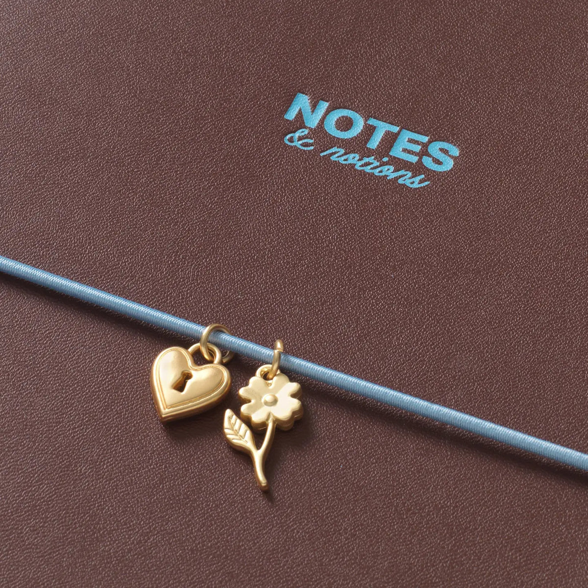 Two gold charms, a heart and a flower with stem and leaf, hanging from a thin blue wire against a textured brown background with the brand name 'NOTES & notions' printed in light blue.
