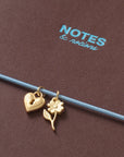 Two gold charms, a heart and a flower with stem and leaf, hanging from a thin blue wire against a textured brown background with the brand name 'NOTES & notions' printed in light blue.