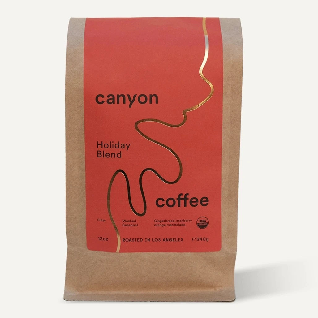 A package of Canyon Coffee's Holiday Blend featuring a minimalist red and kraft paper design with a gold wavy line on the label. The text highlights it as a "12oz" filter coffee roasted in Los Angeles.