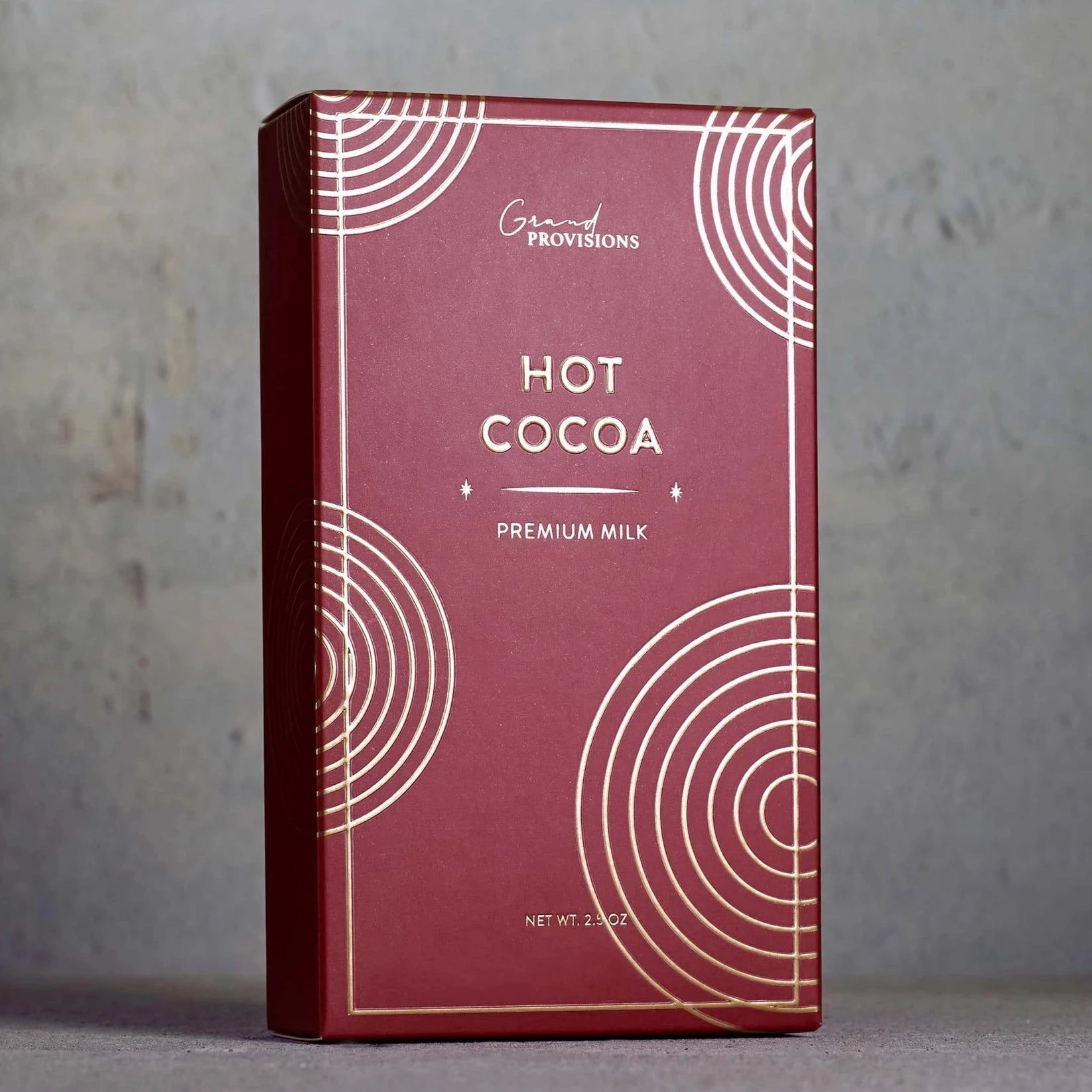 This image displays a premium packaging of hot cocoa labeled "HOT COCOA - Premium Milk" from Grand Provisions. The box has a sleek, minimalistic design with a deep red background and gold circular accents, giving it a sophisticated and festive appearance. The plain gray backdrop enhances the elegant simplicity of the packaging, emphasizing the product’s premium nature.