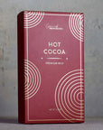 This image displays a premium packaging of hot cocoa labeled "HOT COCOA - Premium Milk" from Grand Provisions. The box has a sleek, minimalistic design with a deep red background and gold circular accents, giving it a sophisticated and festive appearance. The plain gray backdrop enhances the elegant simplicity of the packaging, emphasizing the product’s premium nature.