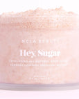 A jar of NCLA Beauty's "Hey, Sugar" exfoliating all-natural body scrub on a soft, light pink background. The scrub has a fluffy, grainy texture visible at the top of the open jar. The frosted jar is labeled with white text showing the product name, description, and size (8.8 oz / 250 g), emphasizing its natural and exfoliating qualities.
