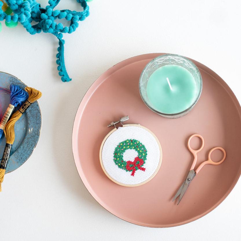 This image shows a creative flat-lay arrangement with a festive touch. At the center is a small embroidery hoop featuring a cross-stitched holiday wreath with green foliage and a red bow. The hoop sits on a pink tray along with peach-handled scissors and a green candle. To the side, a blue plate holds colorful embroidery threads in yellow, blue, and red, along with a spool of red thread. In the top corner, turquoise pom-pom trim adds a playful element.  