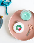 This image shows a creative flat-lay arrangement with a festive touch. At the center is a small embroidery hoop featuring a cross-stitched holiday wreath with green foliage and a red bow. The hoop sits on a pink tray along with peach-handled scissors and a green candle. To the side, a blue plate holds colorful embroidery threads in yellow, blue, and red, along with a spool of red thread. In the top corner, turquoise pom-pom trim adds a playful element.  