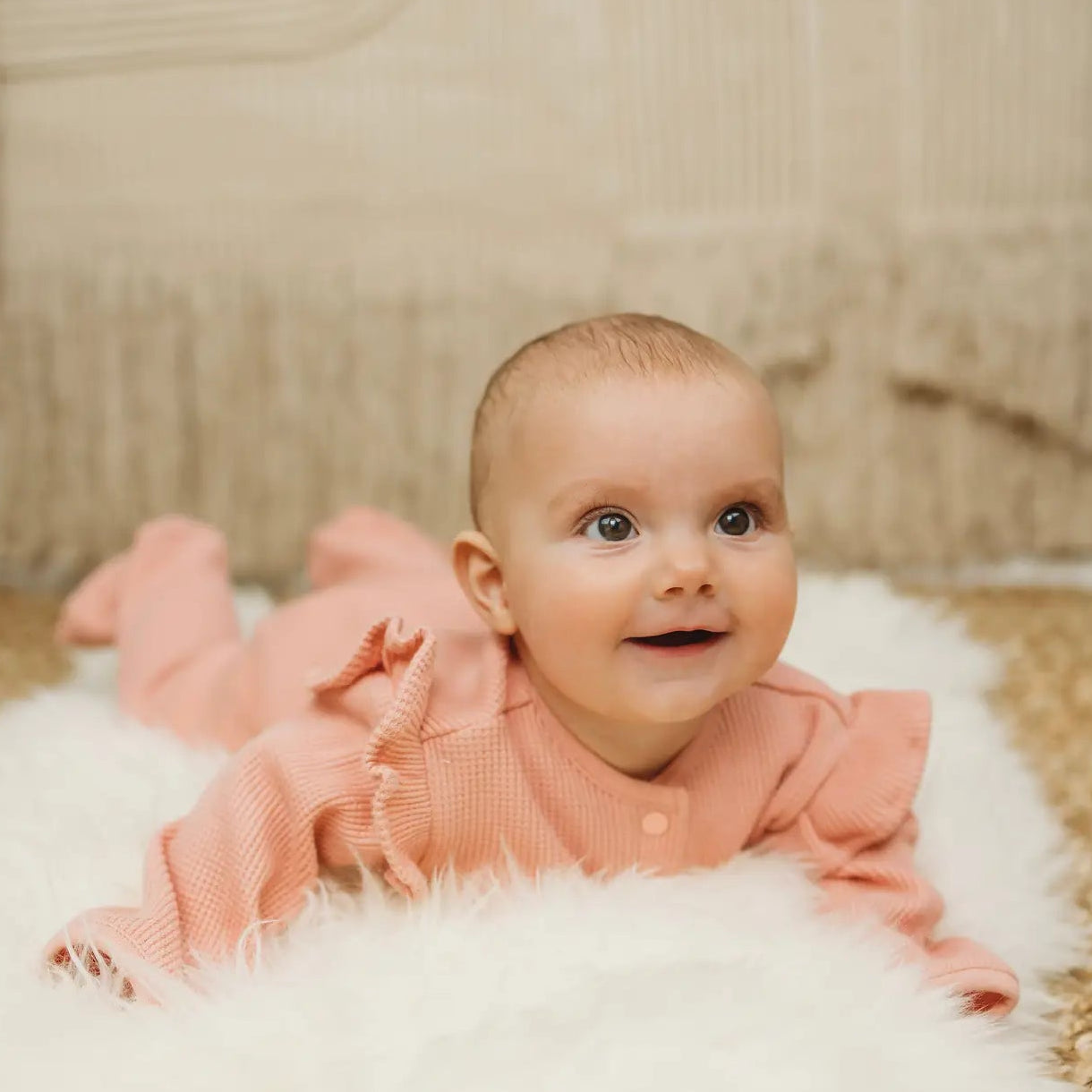 Baby in a pink outfit lying on a fluffy white rug with a wooden floor background