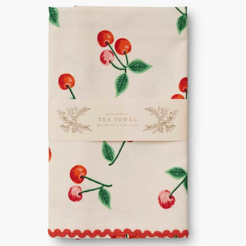 The cherry-patterned tea towel is neatly folded and wrapped in a Rifle Paper Co. branded paper label. The label reads "Tea Towel, 100% Cotton, 21 x 28 inches," showcasing its elegant packaging.

