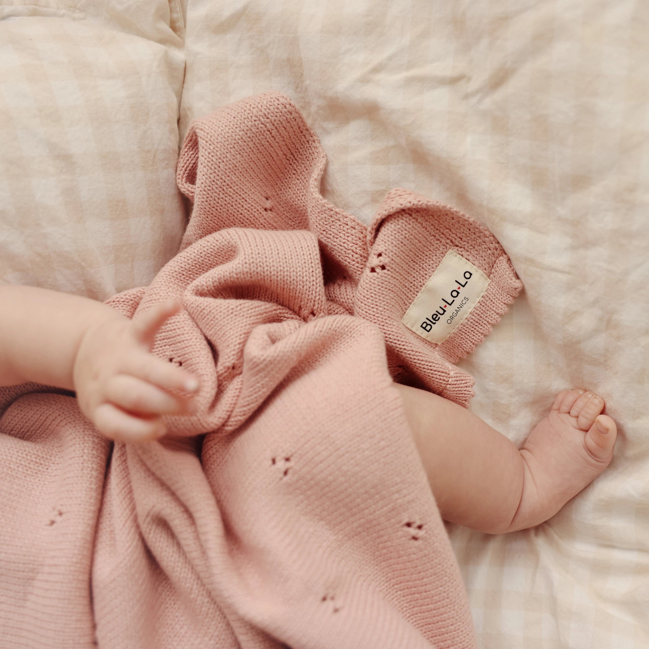 Baby wearing pink knitted booties and holding a pink blanket on a soft surface