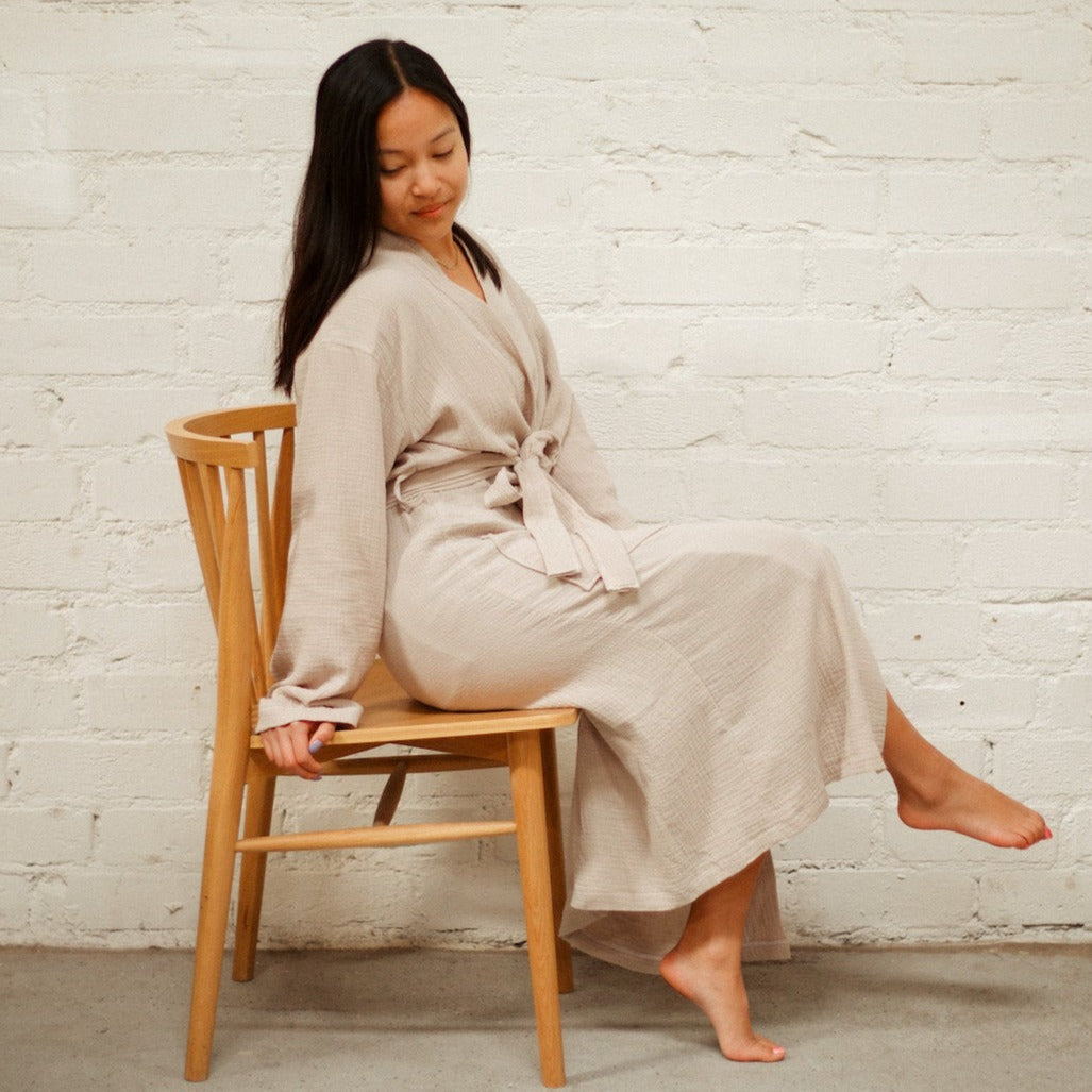 A girl with straight, medium length dark brown hair wearing an oatmeal colored mid-calf length muslin robe, sitting in a brown wooden chair. She is in front of a white brick background.