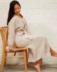 A girl with straight, medium length dark brown hair wearing an oatmeal colored mid-calf length muslin robe, sitting in a brown wooden chair. She is in front of a white brick background.