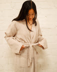 A girl with straight, medium length dark brown hair wearing an oatmeal colored mid-calf length muslin robe. She is standing in front of a white brick background.