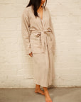 A girl with straight, medium length dark brown hair wearing an oatmeal colored mid-calf length muslin robe. She is standing in front of a white brick background.