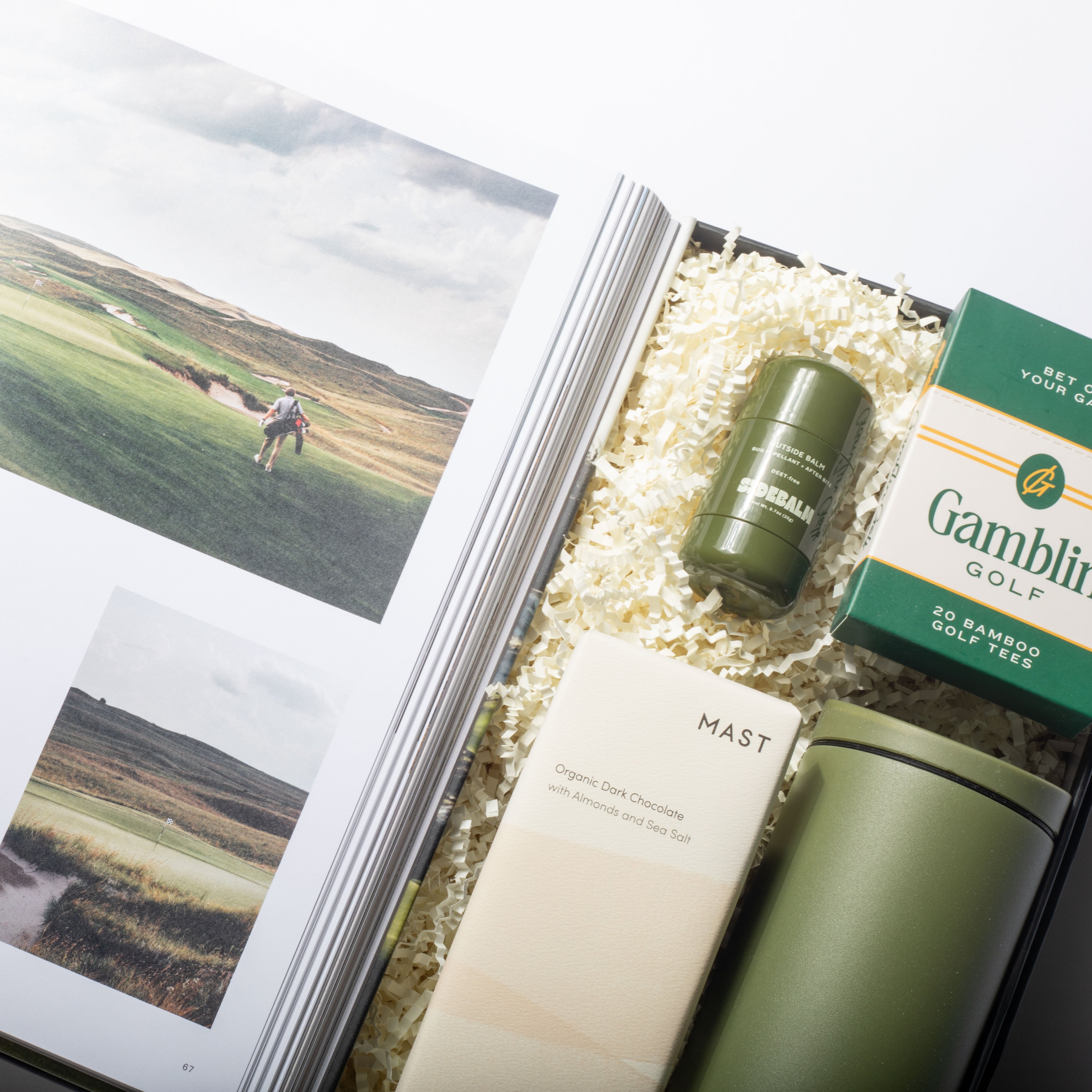 Golf-themed gift set with a magazine, green tumbler, and box of Gamblin Golf tees on a white background.