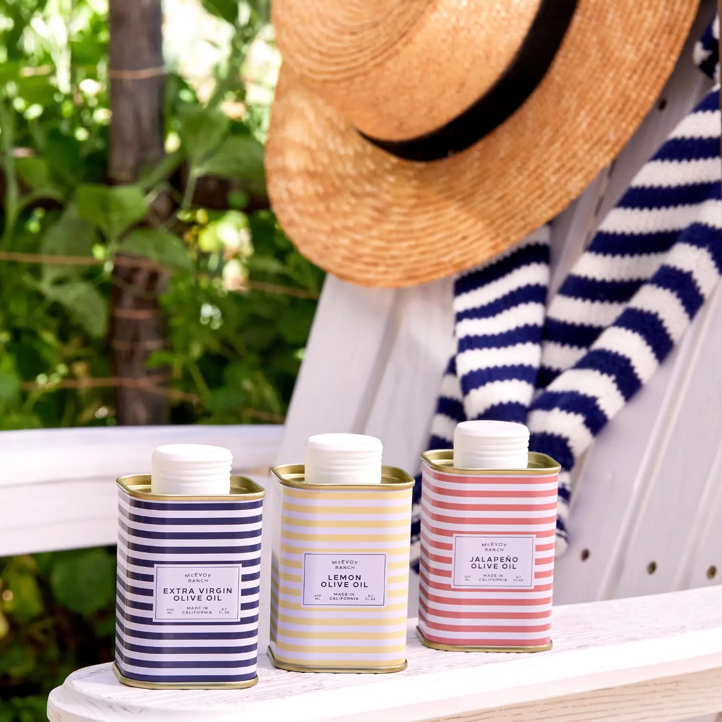 Three striped metal tins of olive oil are displayed on a white wooden table outdoors, with a person wearing a straw hat and a navy-and-white striped sweater partially visible in the background.