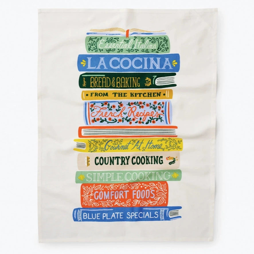 A colorful tea towel featuring a stack of illustrated cookbooks. The books are labeled with titles such as "La Cocina," "Bread & Baking," "Country Cooking," and "Comfort Foods," among others. The artwork has a vibrant and vintage-inspired style.