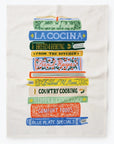 A colorful tea towel featuring a stack of illustrated cookbooks. The books are labeled with titles such as "La Cocina," "Bread & Baking," "Country Cooking," and "Comfort Foods," among others. The artwork has a vibrant and vintage-inspired style.