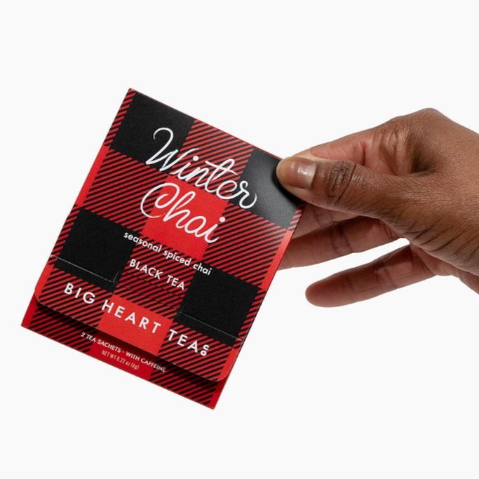 hand holding tea sachet that is a red & black plaid checkered pattern. tea sachet reads Winter Chai in white script, seasonal spiced chai black tea by big heart tea co, on white background. 