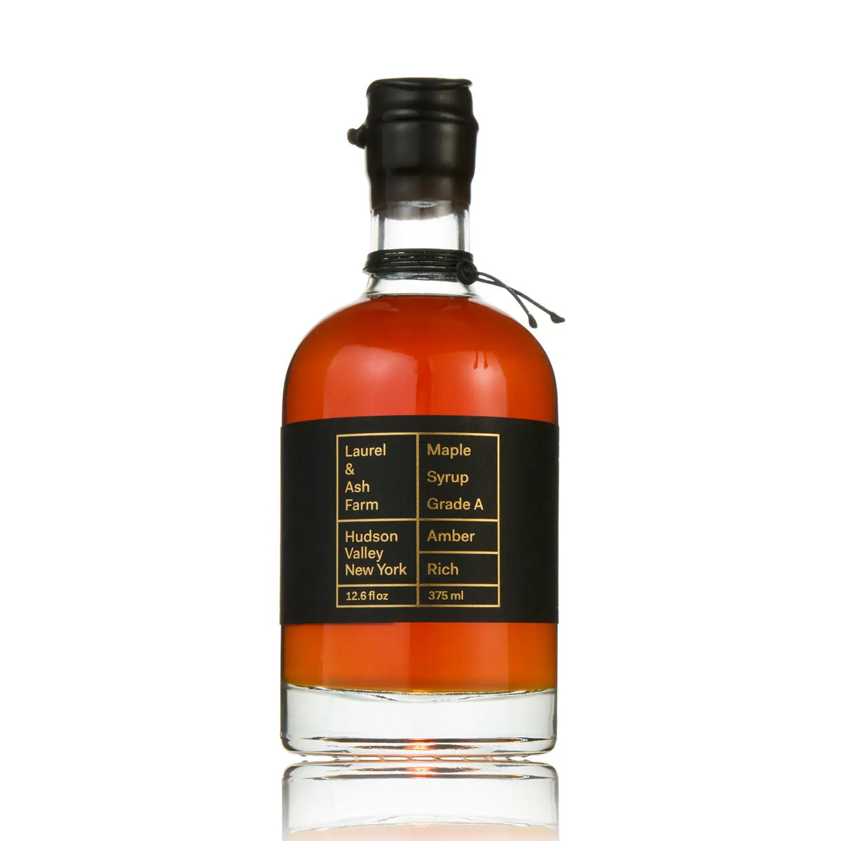 A bottle of Laurel &amp; Ash Farm Maple Syrup, Grade A, Amber flavor, in a clear glass bottle with amber liquid, a black label with gold text, and a black screw cap, shown on a white background with a reflection.