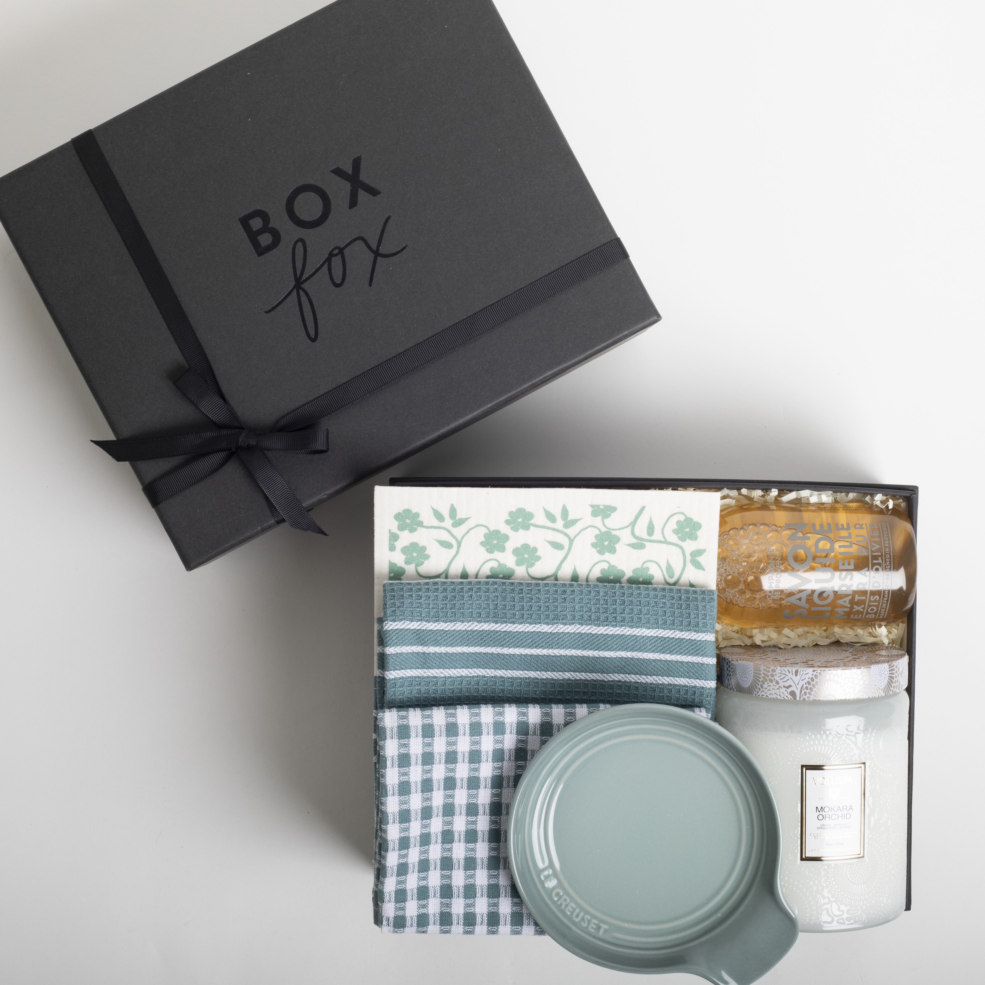 Gift box with 'Box Fox' branding containing folded fabric and a candle on a light gray background