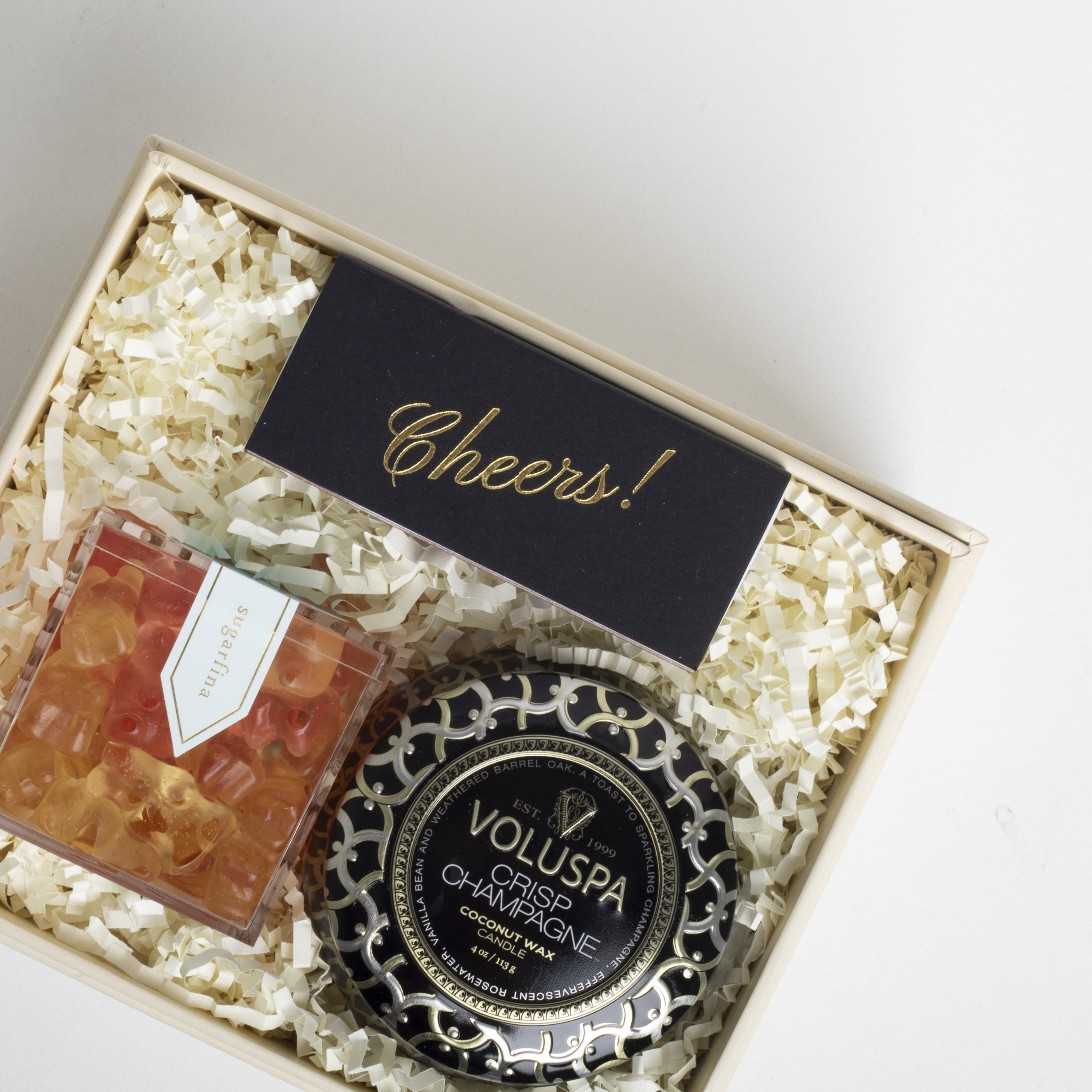 Decorative candle with &#39;Cheers!&#39; card and Voluspa branding in a gift box.