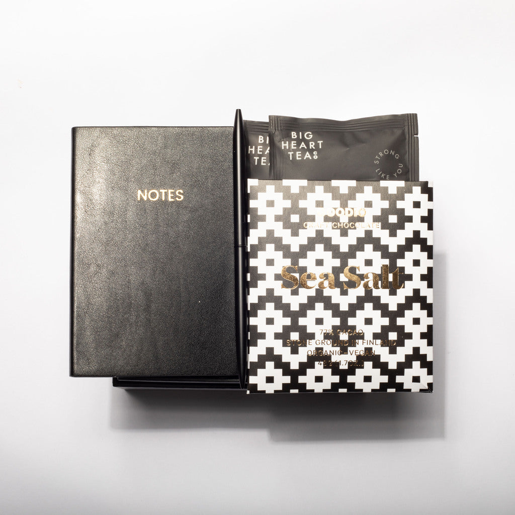 A Sea Salt tea gift set featuring a black textured notebook with gold lettering, a black tea sachet, and a black-and-white geometric patterned box, arranged on a light gray background.