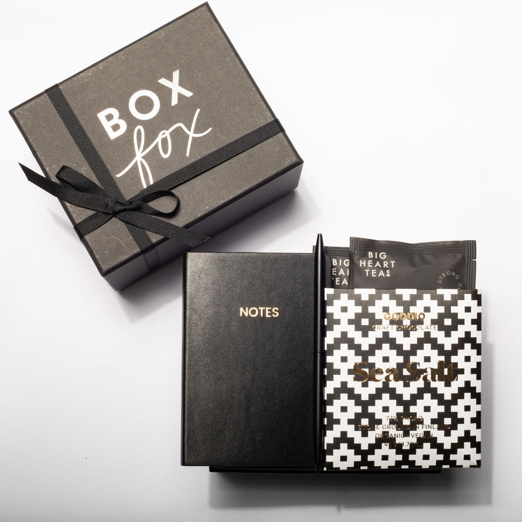A boxed stationery gift set on a white background, including a dark gray gift box labeled 'BOX fox' with a black ribbon, a black notebook with gold 'NOTES' text, and a patterned black-and-white packet labeled 'Sea Salt' and 'Big Heart Tea'.