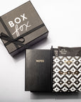 A boxed stationery gift set on a white background, including a dark gray gift box labeled 'BOX fox' with a black ribbon, a black notebook with gold 'NOTES' text, and a patterned black-and-white packet labeled 'Sea Salt' and 'Big Heart Tea'.