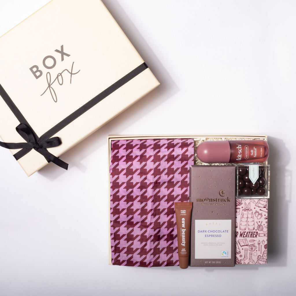 A curated gift box set from Box Fox, featuring a cream-colored gift box with a black ribbon on the left, and an open box on the right containing a pink and purple houndstooth scarf, a lip balm, a tube of espresso scrub, chocolates, and a weathered notebook.