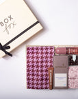 A curated gift box set from Box Fox, featuring a cream-colored gift box with a black ribbon on the left, and an open box on the right containing a pink and purple houndstooth scarf, a lip balm, a tube of espresso scrub, chocolates, and a weathered notebook.