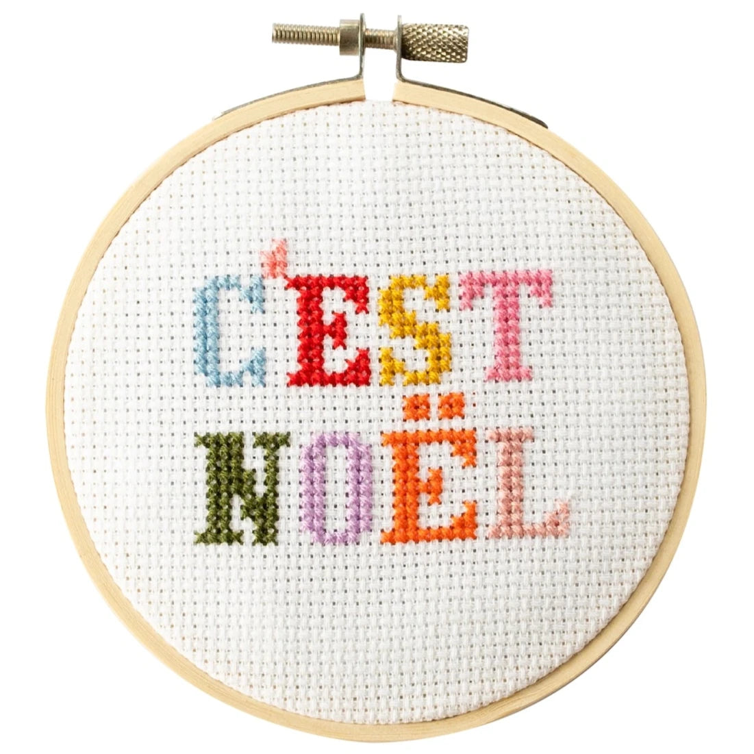 This is an image of a cross-stitch embroidery hoop featuring the French text "C'EST NOËL," which translates to "It's Christmas." The letters are stitched in a variety of bright colors, giving it a festive and playful appearance. The embroidery is set inside a wooden hoop, typically used for holding fabric taut while stitching. 