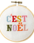 This is an image of a cross-stitch embroidery hoop featuring the French text "C'EST NOËL," which translates to "It's Christmas." The letters are stitched in a variety of bright colors, giving it a festive and playful appearance. The embroidery is set inside a wooden hoop, typically used for holding fabric taut while stitching. 