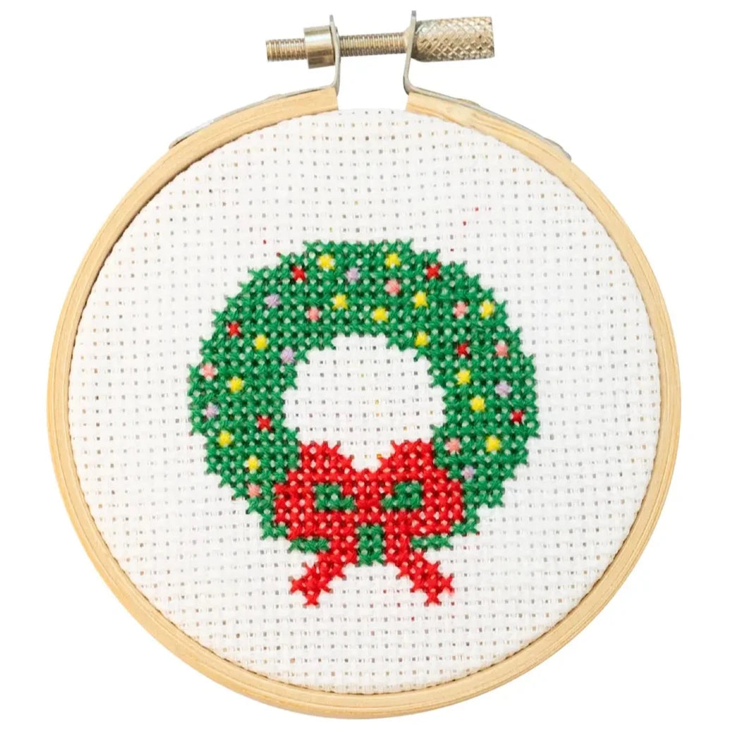 This image features a small embroidery hoop containing a festive cross-stitch design of a holiday wreath. The wreath is made of green stitches, with colorful accents resembling ornaments in red, yellow, and white. A bright red bow decorates the bottom of the wreath, enhancing the seasonal theme. The design is framed neatly within a light wooden hoop, showcasing the clean and minimalist look of a handmade holiday craft.
