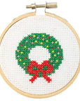 This image features a small embroidery hoop containing a festive cross-stitch design of a holiday wreath. The wreath is made of green stitches, with colorful accents resembling ornaments in red, yellow, and white. A bright red bow decorates the bottom of the wreath, enhancing the seasonal theme. The design is framed neatly within a light wooden hoop, showcasing the clean and minimalist look of a handmade holiday craft.
