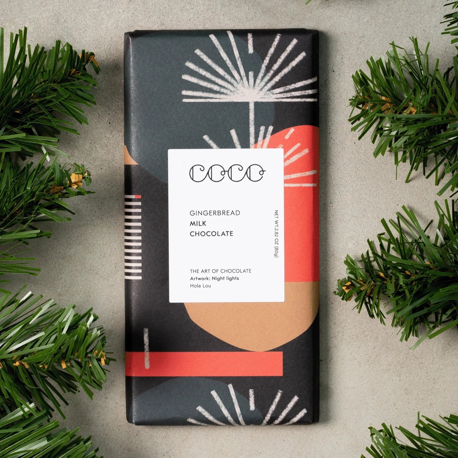 COCO Gingerbread Milk Chocolate Bar with black, red, white and brown packaging on top of a grey background with pine branches around it