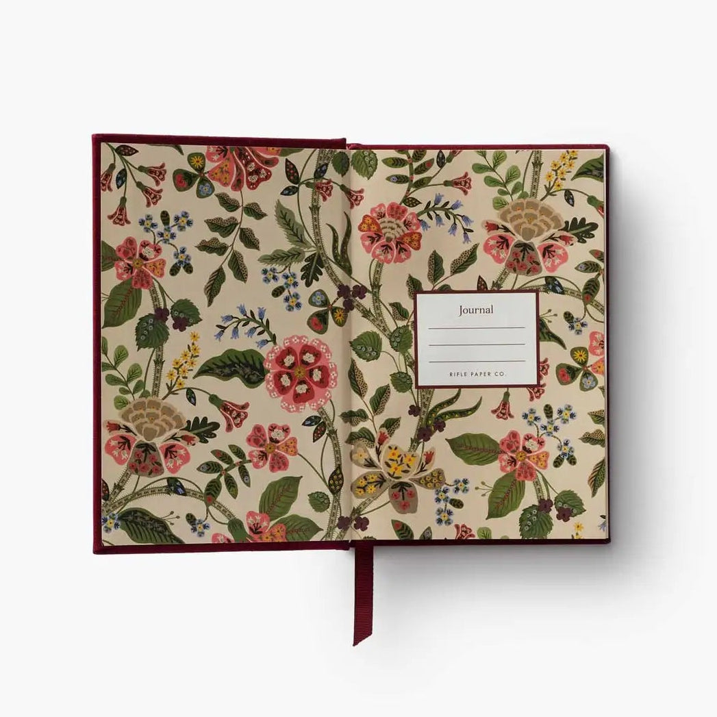 An open floral journal with a cream-colored cover decorated with a dense botanical pattern of pink, red, green, and yellow flowers. It features a dark burgundy cover edge, a matching burgundy ribbon bookmark, and a white label on the right page.