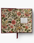 An open floral journal with a cream-colored cover decorated with a dense botanical pattern of pink, red, green, and yellow flowers. It features a dark burgundy cover edge, a matching burgundy ribbon bookmark, and a white label on the right page.
