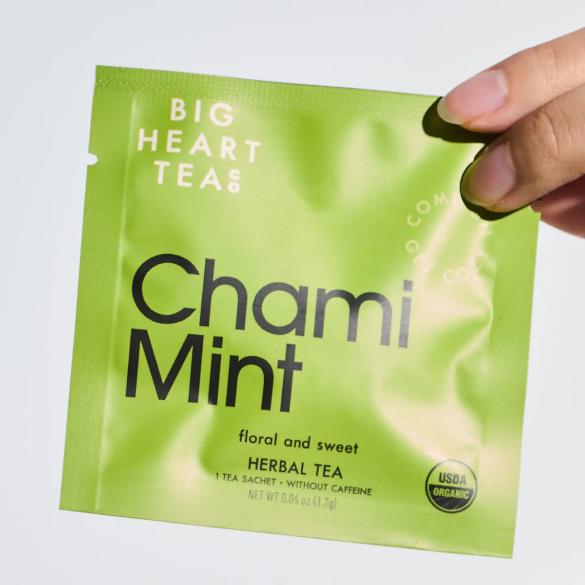 A hand holding a lime green Big Heart Teas Chami Mint herbal tea sachet against a plain white background.