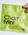 A hand holding a lime green Big Heart Teas Chami Mint herbal tea sachet against a plain white background.