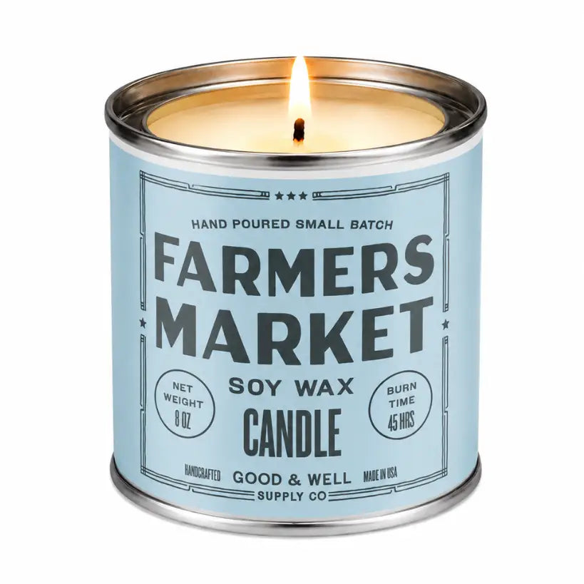 A lit soy wax candle in a silver metal tin with a light blue label reading 'FARMERS MARKET' and 'GOOD &amp; WELL SUPPLY CO' on a white background.