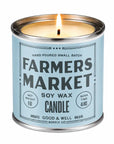 A lit soy wax candle in a silver metal tin with a light blue label reading 'FARMERS MARKET' and 'GOOD & WELL SUPPLY CO' on a white background.