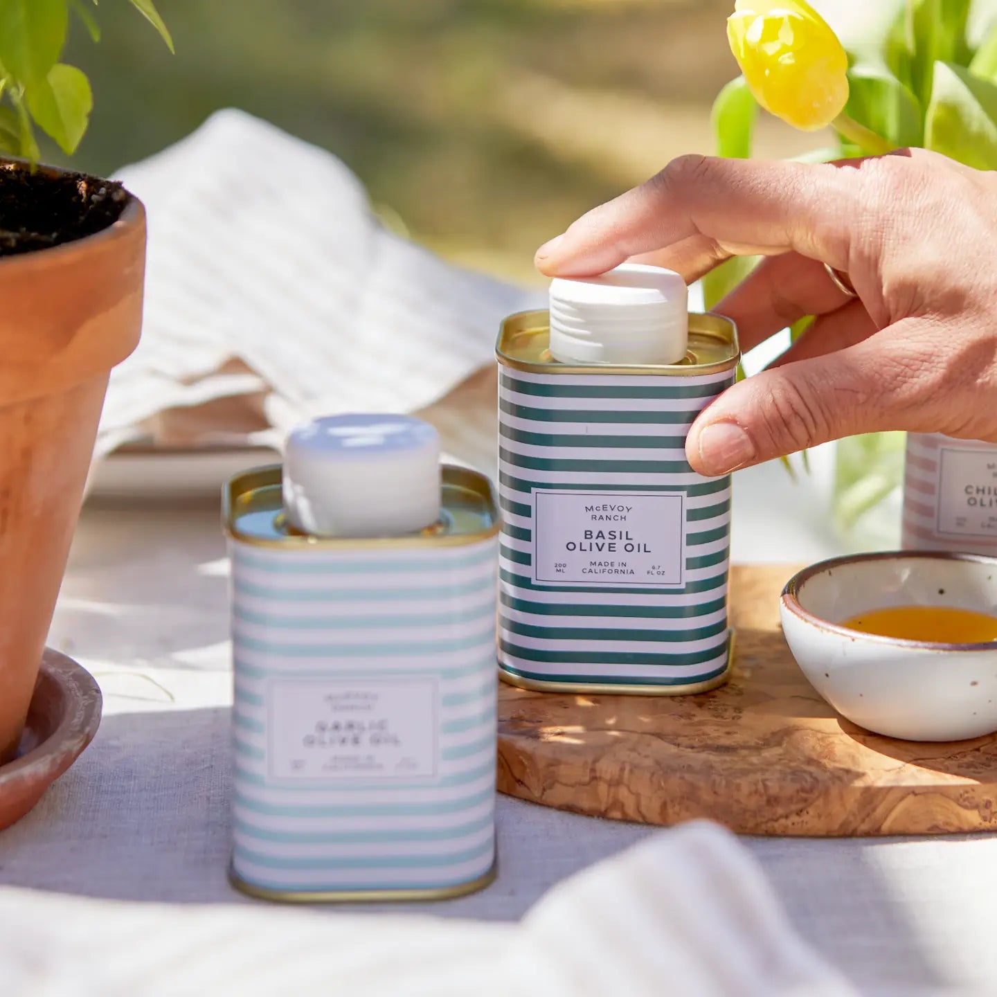 Person opening a striped can of Mevo+ basil olive oil on a table with a blurred outdoor background.