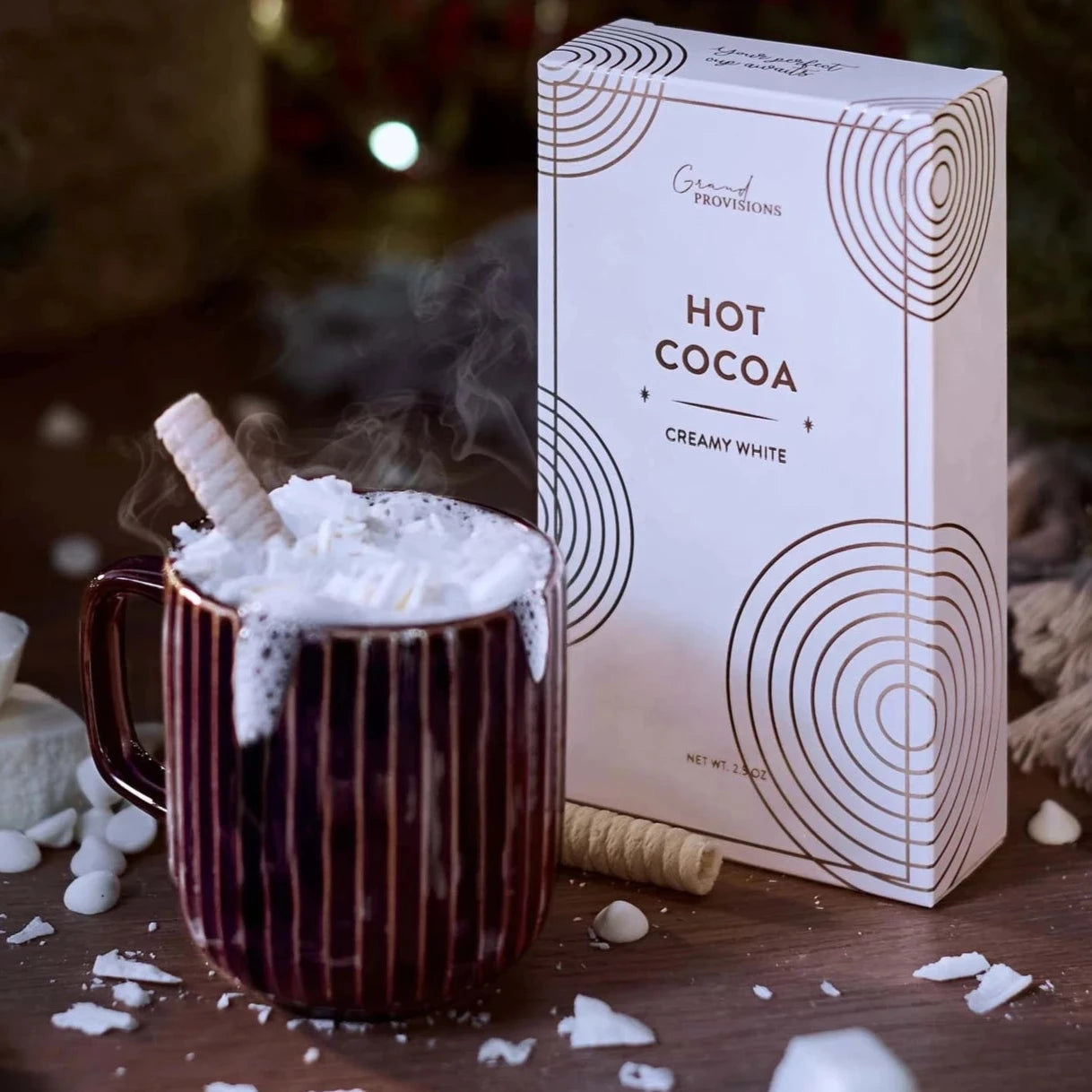 This image showcases a cozy holiday scene with the same "Hot Cocoa" product from Grand Provisions in the "Creamy White" flavor. The box is placed next to a steaming mug of hot cocoa topped with whipped cream, white chocolate shavings, and a wafer stick. The scene is set with festive decorations like fir branches, twinkling lights, and scattered marshmallows, creating a warm, wintery ambiance.