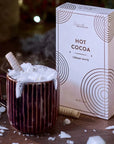 This image showcases a cozy holiday scene with the same "Hot Cocoa" product from Grand Provisions in the "Creamy White" flavor. The box is placed next to a steaming mug of hot cocoa topped with whipped cream, white chocolate shavings, and a wafer stick. The scene is set with festive decorations like fir branches, twinkling lights, and scattered marshmallows, creating a warm, wintery ambiance.