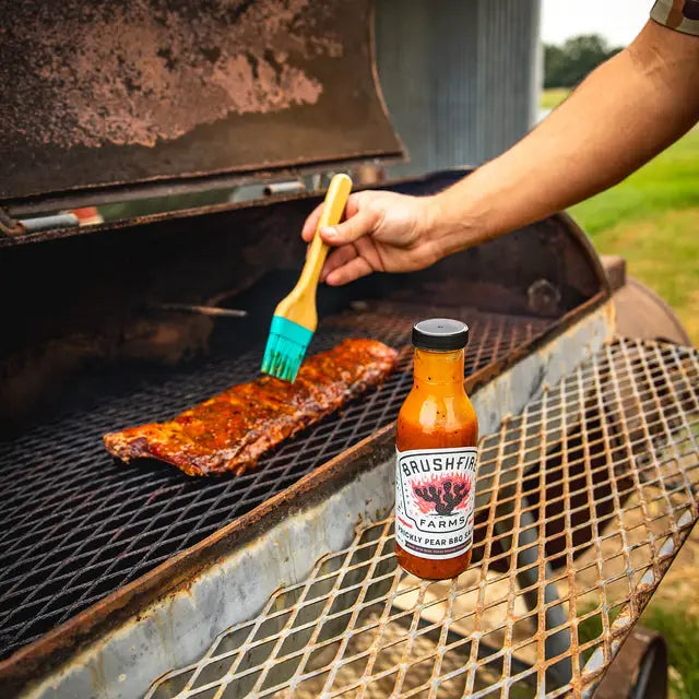 Person brushing ribs with a brush on a grill next to a bottle of Brushfire Farms sauce.