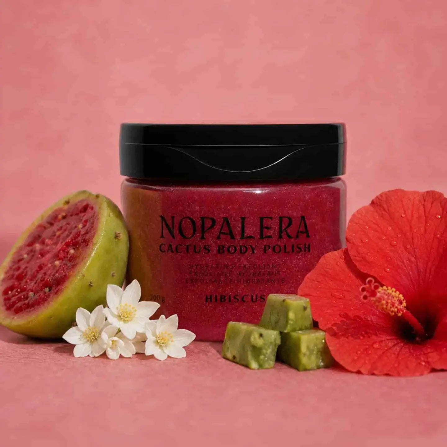 A jar of Nopalera Cactus Body Polish in Hibiscus scent, styled with a halved green guava, white jasmine flowers, green cactus pieces, and a red hibiscus flower on a pink background.