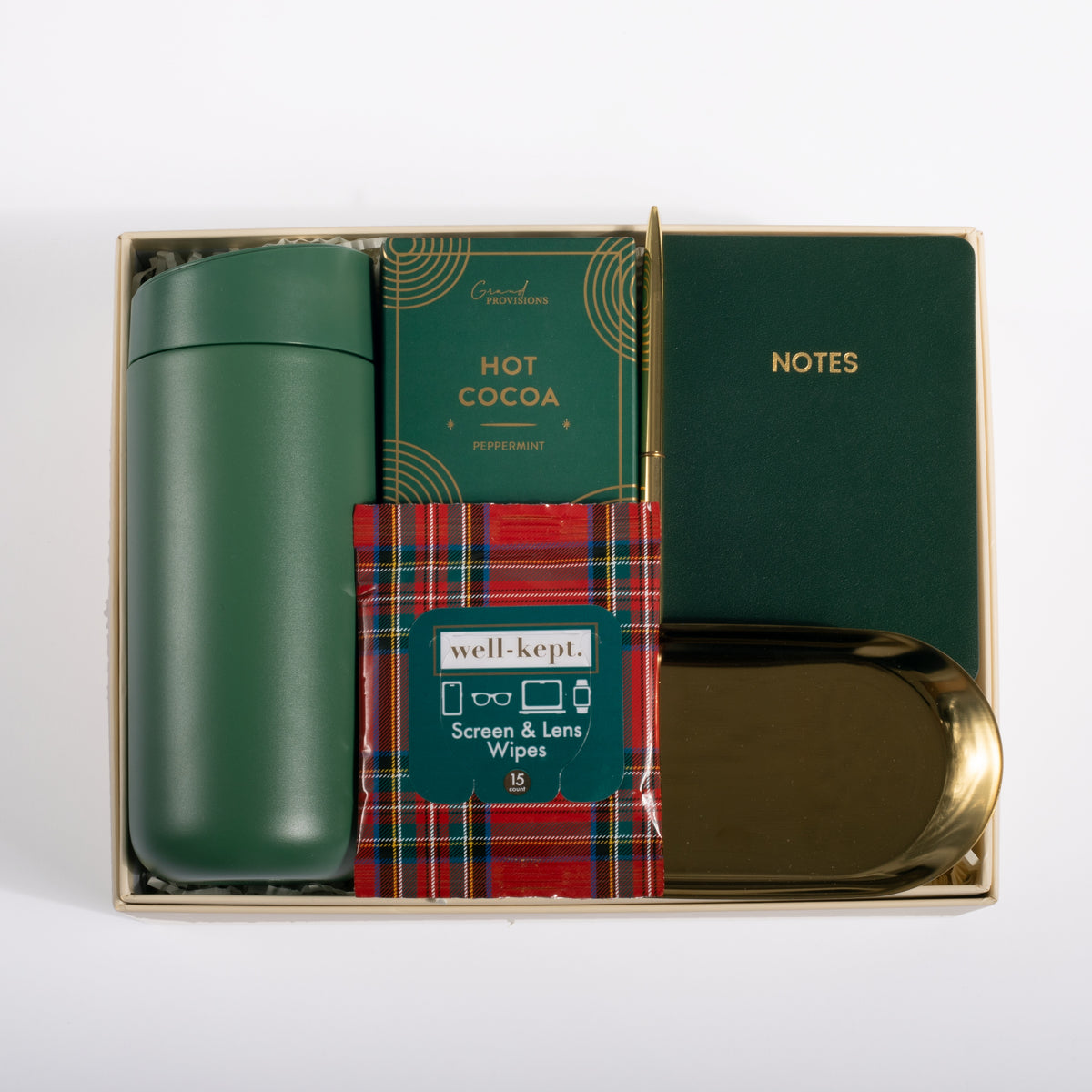 A curated gift set in a gold-toned box containing a dark green insulated bottle, a green 'HOT COCOA' card, a red tartan 'well-kept.' screen and lens wipes packet, a dark green 'NOTES' notebook, and a gold-toned tray, all arranged on a white background.
