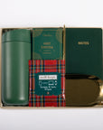 A curated gift set in a gold-toned box containing a dark green insulated bottle, a green 'HOT COCOA' card, a red tartan 'well-kept.' screen and lens wipes packet, a dark green 'NOTES' notebook, and a gold-toned tray, all arranged on a white background.
